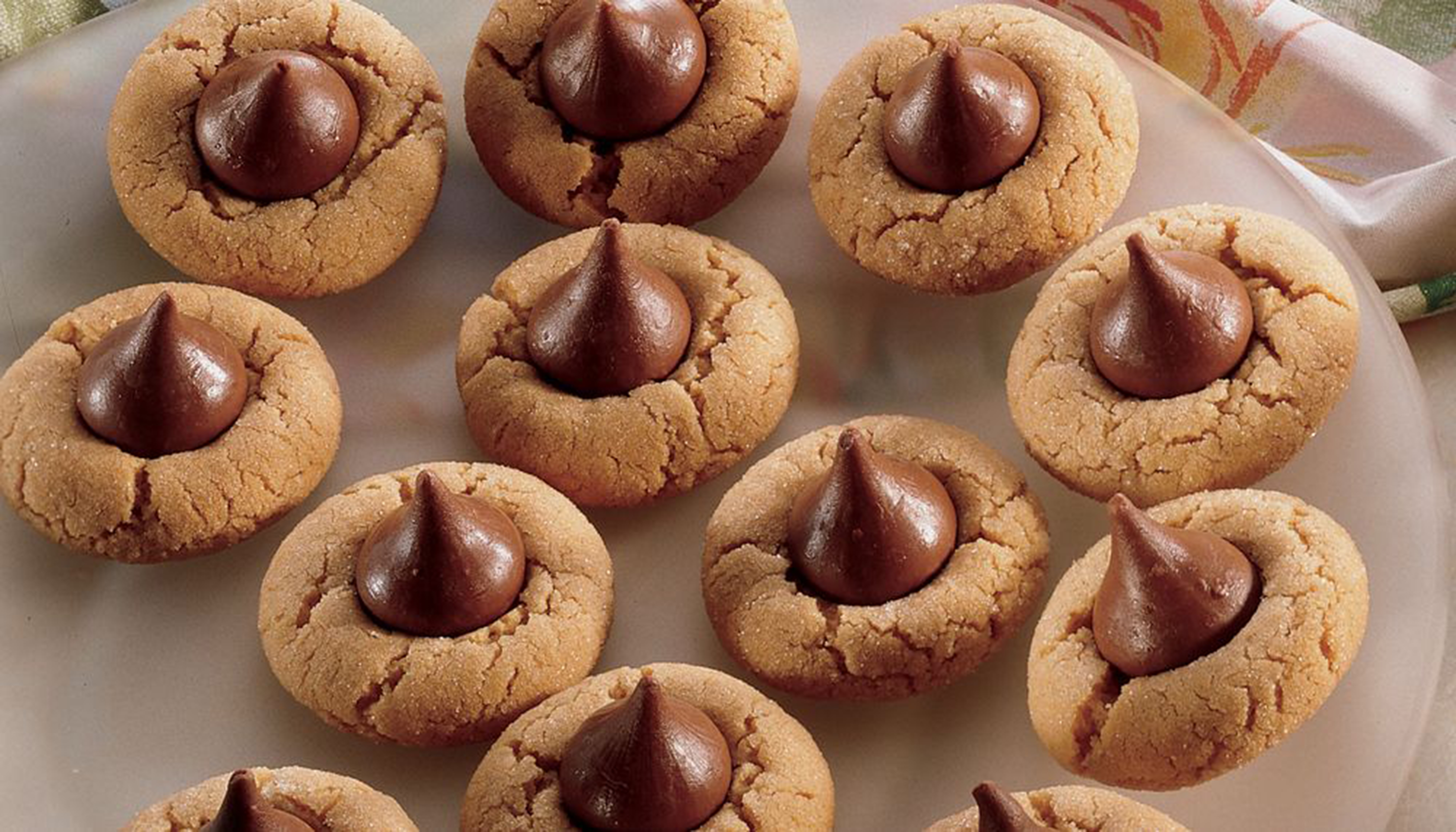 A close-up photograph of a platter of classic peanut butter blossom cookies. Each round, golden-brown cookie has a signature cracked surface and a milk chocolate kiss candy pressed into the center.