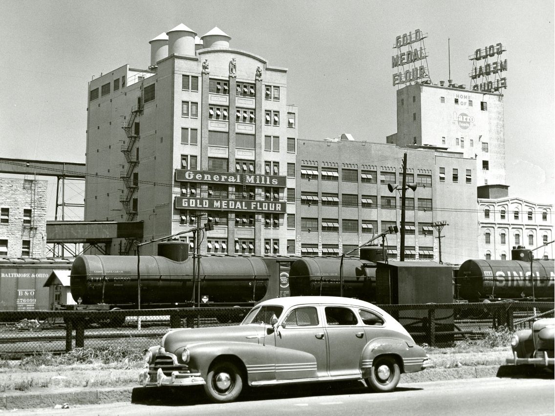 A black and white photograph shows the General Mills building with "General Mills" and "Gold Medal Flour" signs prominently displayed. On the roof of a taller building behind it, large signs spell out "GOLD MEDAL FLOUR." In the foreground, a vintage car is parked on the side of a road, and behind it, several train cars, including tank cars and a boxcar are visible on tracks.